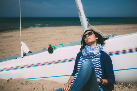 Portrait of brunette in sunglasses leaning on boat on beach in sunlightの写真素材