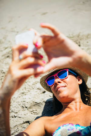 Portrait of smiling adult woman in hat and sunglasses taking self-portrait via cell phoneの写真素材