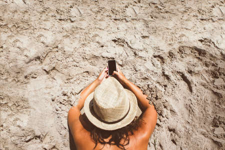 Top view of unrecognizable woman in hat lying on beach and using smartphone.の写真素材