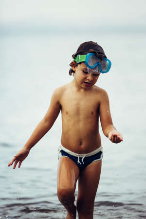 Portrait of adorable boy walking in water with mask on head and holding something in handの写真素材