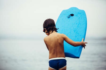 Rear view of little boy in trunks and goggles holding blue bodyboard in seaの写真素材