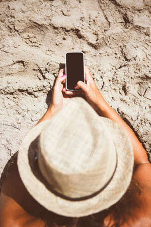 Overhead of unrecognizable woman in hat chatting over smartphone on beach in sunlight