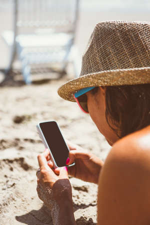 Overshoulder view of adult woman on beach using smartphone in sunlightの写真素材