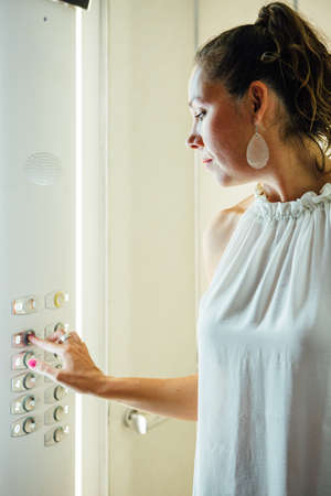 Side view of adult woman in white blouse with ponytail pushing button with finger inside elevator