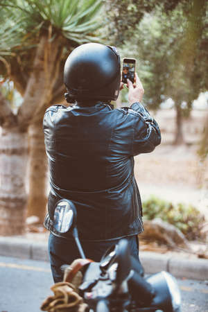 Back view of man in leather jacket and bike helmet taking self-portrait via phoneの写真素材