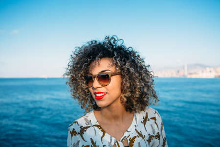 Portrait of smiling pretty woman with curly hair in sunglasses looking away. Ocean on background.
