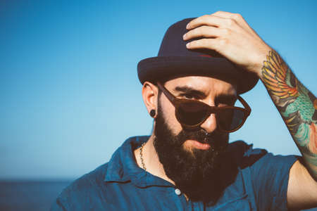 Portrait of stylish bearded man with colorful tattoo on hand lookig over sunglasses and holding hatの写真素材