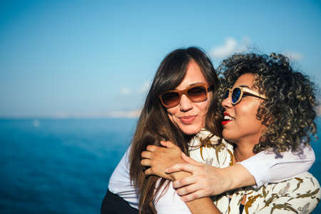 Mixed race girl with curly hair and pretty brunette hugs against seascape and blue skyの写真素材