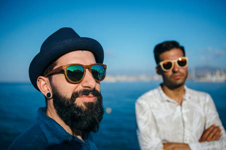 Portrait of two hipsters in sunglasses and hat posing against oceanの写真素材
