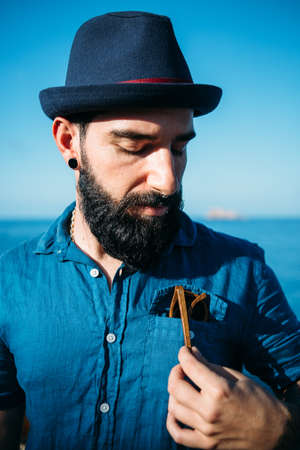 Portrait of stylish bearded man with blue hat takes sunglasses from pocket against ocean.の写真素材