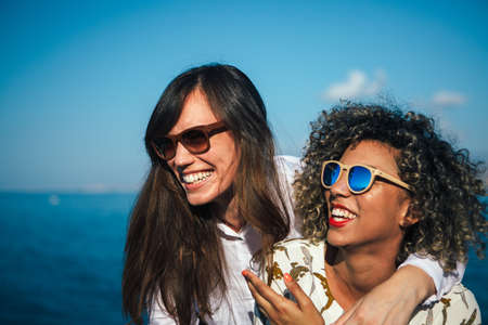 Mixed race girl with curly hair and pretty brunette hugs and laughing against seascape and blue skyの写真素材