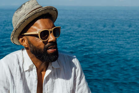 Portrait of african stylish bearded man in sunglasses and straw hat against oceanの写真素材