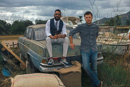 Portrait of two adult stylish men posing at rusty retro car in countrysideの写真素材