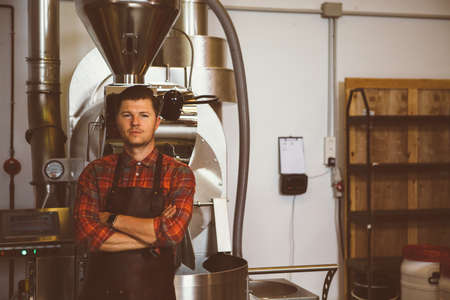 Portrait of adult handsome man with arms crossed in apron against of roasting coffee machineの写真素材
