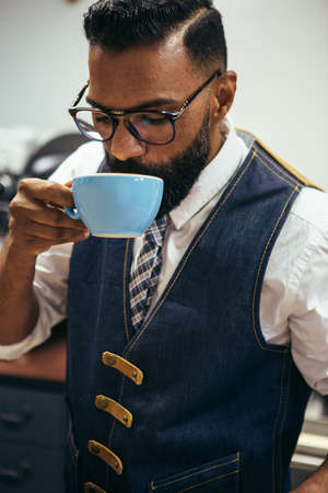 Portrait of bearded black man in glasses and waistcoat drinking coffee from blue cupの写真素材