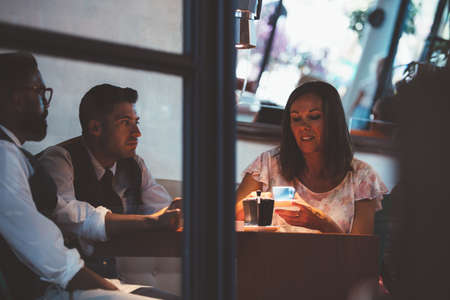 Adult woman and two men sitting at table with coffee in coffee shop.View through the doors.