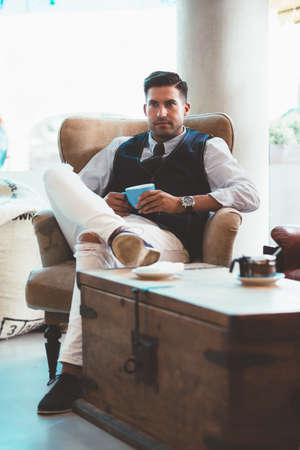 Portrait of good-looking man with blue cup sitting on arm-chair
