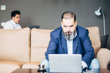 Portrait of bearded businessman with pen in mouth working on laptop against of young manの写真素材