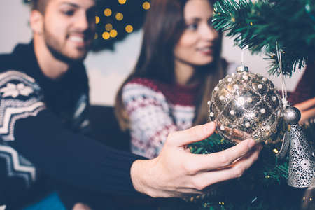 Close up hand of man with girl in living room hanging golden bauble on Christmas tree.