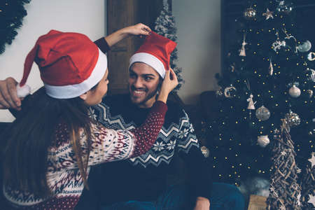 Laughing loving couple in sweaters trying on Santa hats in room decorated to Christmas. の写真素材