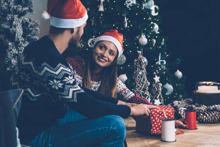 Young couple wearing Santa hats sitting on sofa and decorating Christmas gift box together. の写真素材