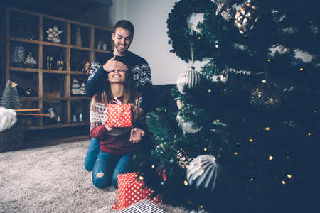 Man closing eyes to woman holding present and smiling in room decorated to Christmas. の写真素材
