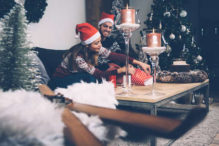 Laughing couple in sweaters and Santa hats wrapping Christmas present on table with candles and wreath. の写真素材