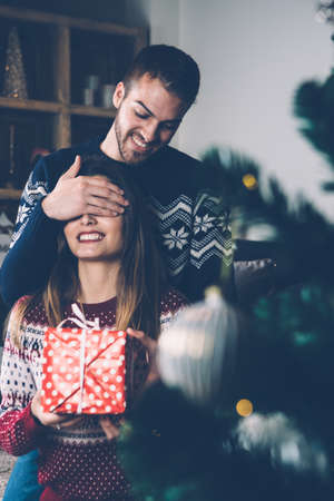 Man making surprising present to girlfriend closing her eyes and laughing in room decorated to Christmas. の写真素材