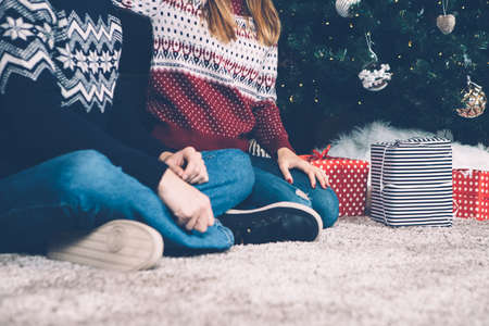 Crop faceless couple in sweaters sitting on carpet near decorated fir tree with presents below. 