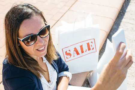 Young smiley female in sunglasses sitting outdoors with packages and taking selfie. の写真素材