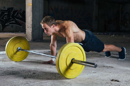 Side view of young man doing push-ups near barbell in abandoned building.の写真素材