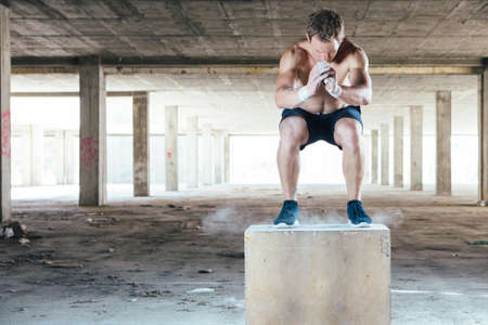 Front view of young muscular man with naked torso doing box jumps in abandoned building.の写真素材