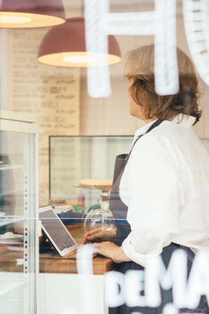 Side view of adult confectioner with tablet at counter. View from behind windowの写真素材