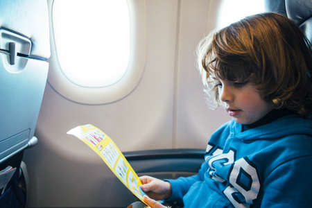 Closeup of a cute blonde boy is reading the safety airplane card while is sitting near te window at the airplane の写真素材