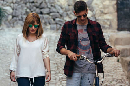 Young man and woman in casual outfits and sunglasses walking down road with bicycle. 