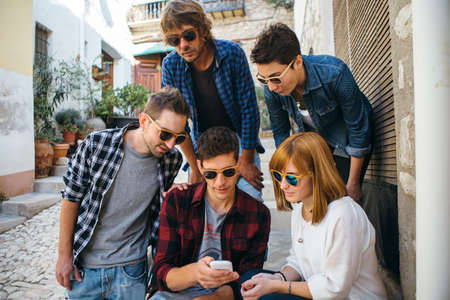 Group of stylish friends in sunglasses using smartphone while sitting outdoors.の写真素材