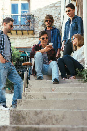 Young trendy friends in sunglasses sitting on stairs and watching smartphone.の写真素材