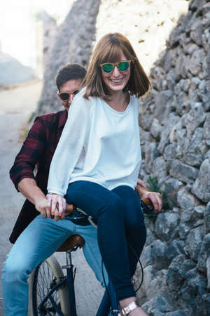 Cheerful young couple in sunglasses riding one bike on narrow old street.の写真素材