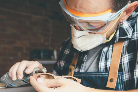 Young artisan in protective eyewear and mask polishing wooden rim for glasses.の写真素材