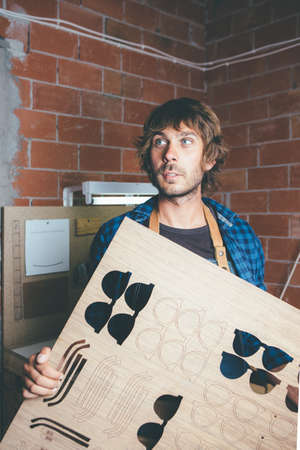 Young bearded worker holding wooden blank for eyeglass frames at workshop.の写真素材