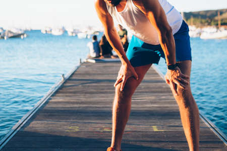 Horizontal outdoors shot of unrecognizable fit man leaning on knees resting on a pier.の写真素材