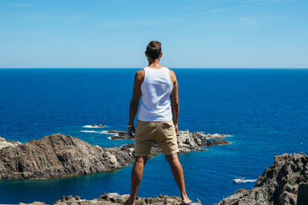 Back view of a muscular man admiring calm blue seascape from rocky shore.の写真素材