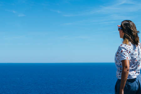 Back view of a woman standing and looking in blue calm sea covering eyes from sun.の写真素材