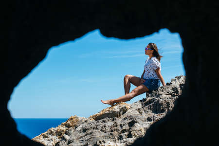 Side view through cave of woman sitting on coastal stone.の写真素材