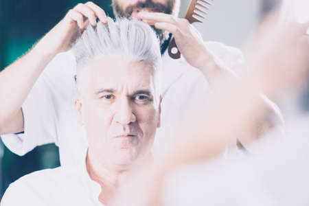 Horizontal indoors shot of adult man sitting in barbershop and looking at reflection.の写真素材