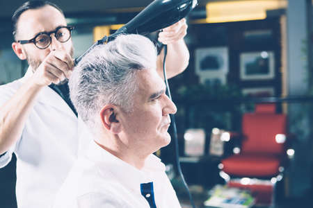 Side view of a stylistworking with drier and comb making a hairdo to mature man.