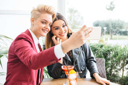 Young stylish girlfriends taking selfie while sitting with drinks at table in cafe.の写真素材