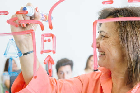 Woman painting scheme on glass board while working with students in classroom.