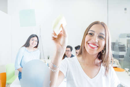 Young content woman posing with sticker behind glass wall of office with coworkers on background.
