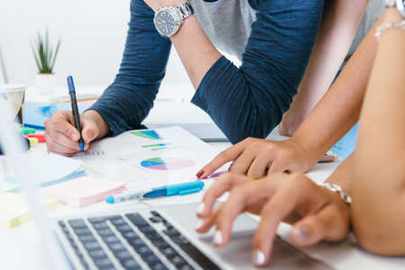 Crop shot of young people sitting at table with laptop and papers and coworking on diagram.